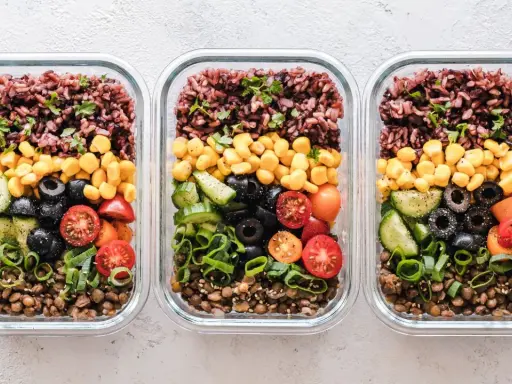 Drei Glasbehälter mit einem gesunden Essensplan für Fitness: Linsen, Gurken, Oliven, Tomaten, Mais und brauner Reis.