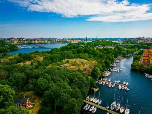 En vacker promenad i Stockholm med grönskande träd, en bro över vattnet och många segelbåtar i hamnen.