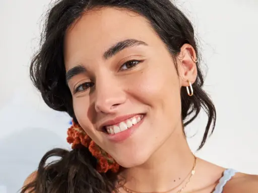 Primo piano di una giovane donna con un sorriso radioso, la sua fronte lucida riflette la luce. Indossa orecchini a cerchio dorati e una collana sottile.