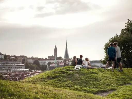 Vänner njuter av utsikten över Haga, Göteborg. En grupp unga människor sitter på en gräskulle med staden i bakgrunden.