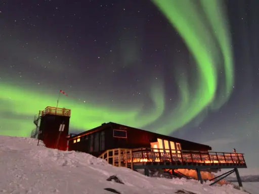 Norrsken dansar över en snöig nationalpark i Lappland. En byggnad med upplysta fönster och en tornliknande struktur står på en sluttning.