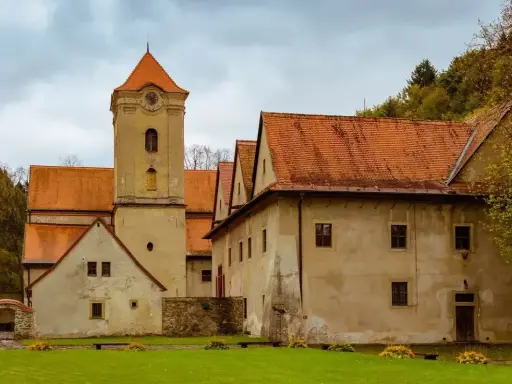 Czerwony klasztor w Pieninach, z charakterystyczną wieżą zegarową i pomarańczowymi dachami, otoczony zielonym trawnikiem i drzewami.