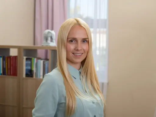 Uśmiechnięta blondynka w jasnej koszuli, za nią regały z książkami.