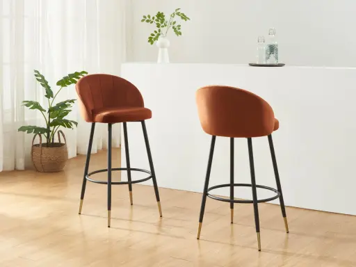 Due poltrone sgabello color ruggine, con gambe nere e dettagli dorati, in un ambiente moderno con piante e una barra.