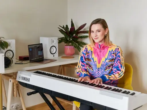 Kobieta gra na pianinie Yamaha. W tle laptop i głośniki, idealne do nauki śpiewu dla dzieci.