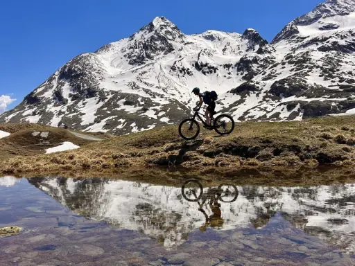 Mountainbiker auf einem epischen Alpencross-Abenteuer, die schneebedeckten Gipfel spiegeln sich in einem Bergsee.
