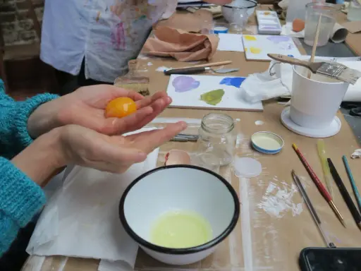 Préparation d'une tempera peinture à l'oeuf : un jaune d'œuf est tenu dans une main au-dessus d'un bol contenant le blanc.