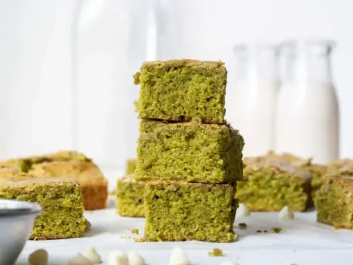 Grüne, saftige matcha brownies, gestapelt und auf einem Tisch arrangiert, mit weißen Schokostückchen und Milchflaschen im Hintergrund.