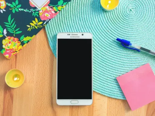 Un smartphone prêt à vous aider à vous organiser au quotidien. Un carnet fleuri, des bougies, un stylo et un post-it rose complètent la scène.