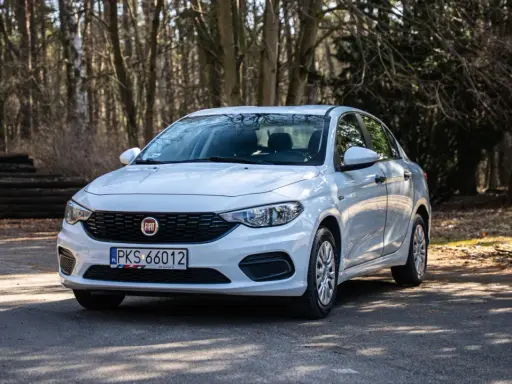 Biały Fiat Tipo 1.4 zaparkowany na leśnej drodze, otoczony drzewami i stertą drewna.