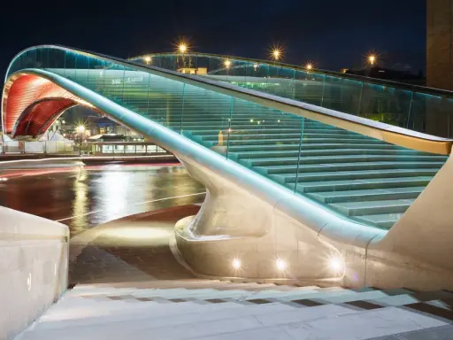 Le pont de la Constitution, illuminé la nuit, avec ses escaliers de verre et ses arches rouges, surplombe un canal aux reflets lumineux.