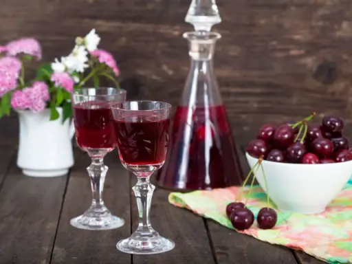 Dwa kieliszki z rubinową nalewką z czereśni na bimbrze, obok miseczka świeżych owoców i bukiecik kwiatów.