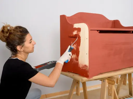 Mujer con guantes blancos pinta con rodillo una caja de madera de color terracota.