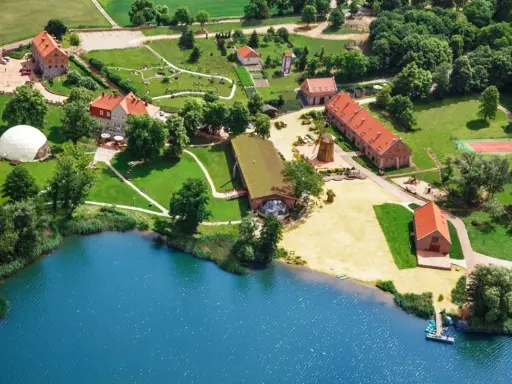 Widok z lotu ptaka na ośrodek SPA nad jeziorem z budynkami, plażą i kortem tenisowym.