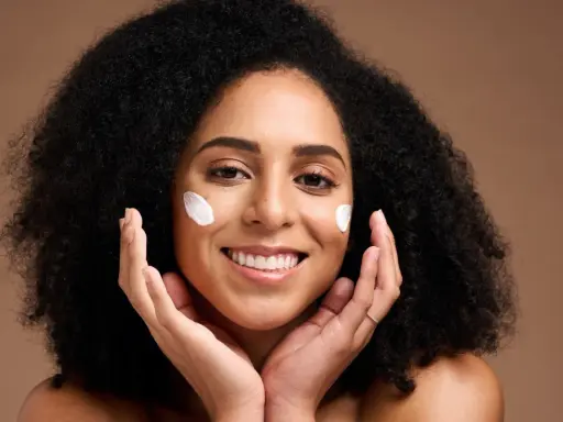 Une femme souriante applique du beurre de karité visage sur ses joues. Ses mains caressent doucement sa peau.