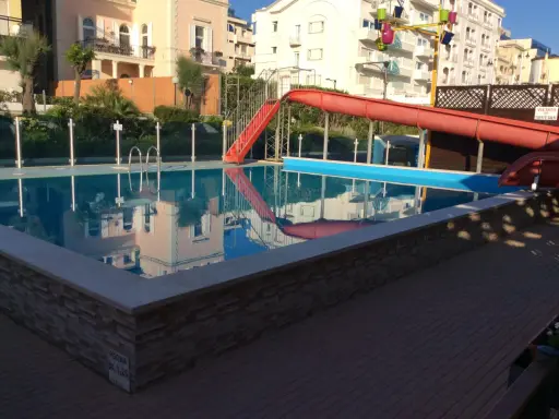 Piscina con scivolo rosso e giochi d'acqua ai Bagni Carmen Cattolica. Riflessi degli edifici nell'acqua azzurra.