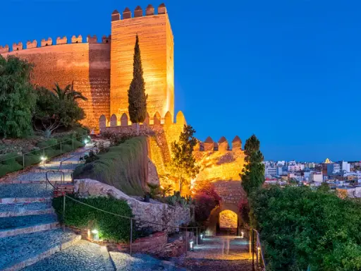 Die Alcazaba von Almería erstrahlt im Abendlicht. Beleuchtete Wege führen durch Gärten zur imposanten Festung.