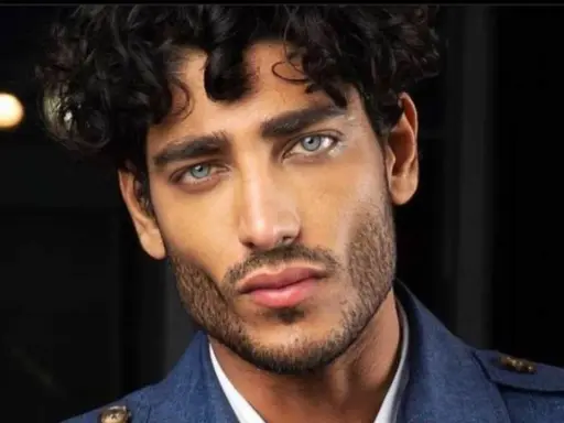 Primo piano di un uomo con splendidi capelli ricci scuri e occhi azzurri penetranti, indossa una giacca blu.