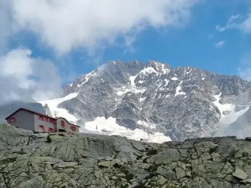Rifugio Gherardi, un edificio con finestre rosse, si erge su un pendio roccioso sotto un imponente massiccio montuoso innevato.