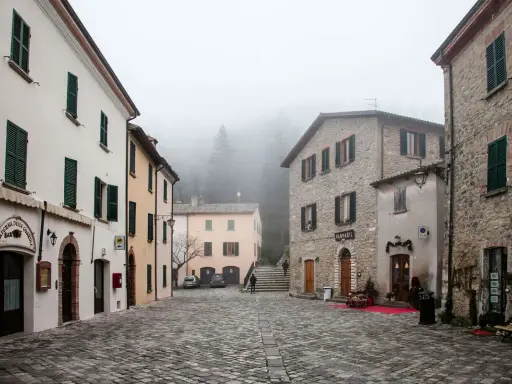 Piazza di San Leo avvolta nella nebbia, con edifici in pietra e intonaco, un bar e una farmacia.