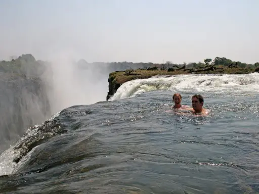 Zwei Personen schwimmen im Devils Pool am Rande der Victoriafälle, mit Gischt und üppiger Vegetation im Hintergrund.