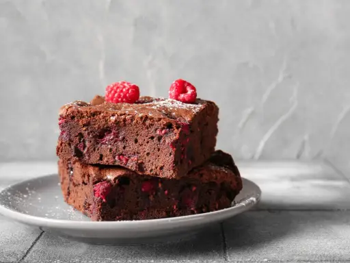 Dwa pyszne brownie z malinami, posypane cukrem pudrem, ułożone na szarym talerzyku.