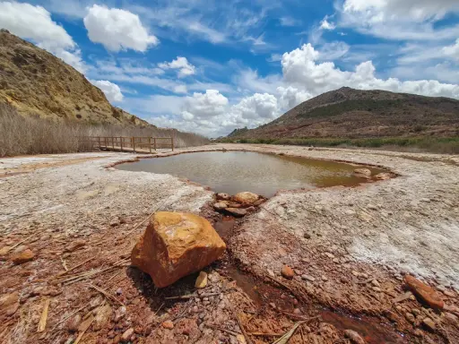Un estanque natural en Les Salinetes de Novelda, rodeado de rocas y vegetación seca, bajo un cielo azul con nubes blancas.
