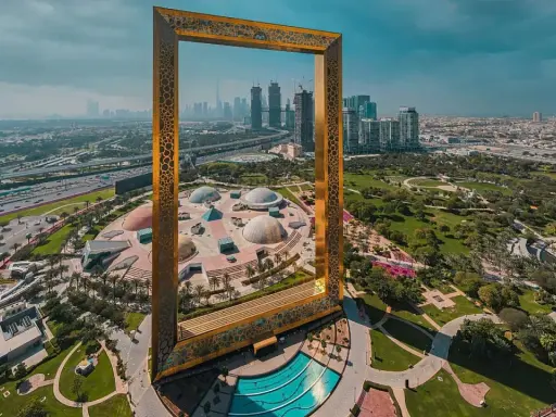 Dubai Frame rahmt die Skyline und die futuristischen Dubai Projekte ein. Ein Park mit Kuppeln und ein Pool sind im Vordergrund.