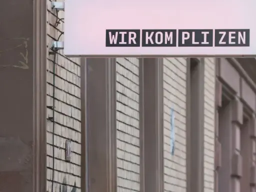 Leuchtreklame mit dem Schriftzug "WIR KOMPLIZEN" über einer Fassade.