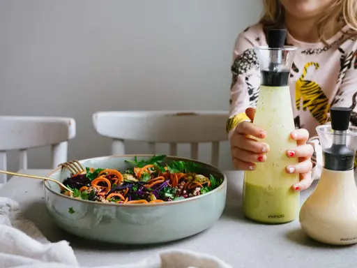 En skål med färgglad sallad och två flaskor dressing till sallad. En flicka håller i den ena flaskan.