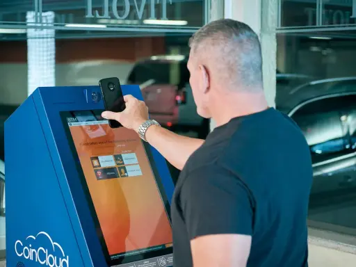 Mężczyzna używa bitomatu CoinCloud do zakupu kryptowalut. Na ekranie widnieje informacja o prowizji i limitach transakcji.