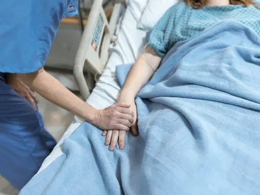Pielęgniarka trzyma za rękę pacjentkę leżącą w łóżku szpitalnym pod niebieskim kocem.