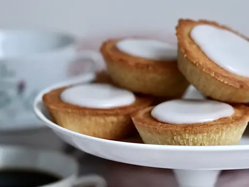 Små, gyllene mazariner med vit glasyr på ett tårtfat. Perfekt till fikat, här är ett enkelt mazariner recept.