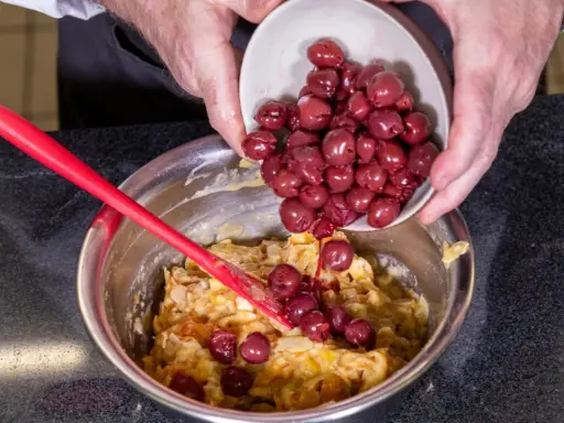 Hände geben Kirschen in eine Schüssel mit Teig für ein einfaches Kirschenmichel Rezept. Ein roter Teigschaber liegt daneben.