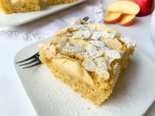 Ein Stück saftiger Apfelkuchen mit Apfelmus, bestreut mit Puderzucker und Mandelblättchen, wartet auf dem Teller.