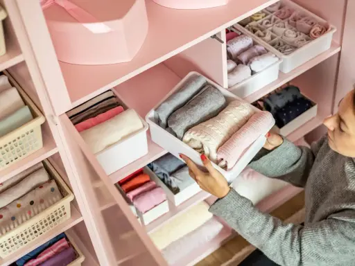 Frau sortiert Kleidung in einem rosafarbenen Schrank. Die Fächer sind ordentlich mit gefalteten Kleidungsstücken gefüllt, die nach Farben sortiert sind, um eine harmonische Reihenfolge zu schaffen.