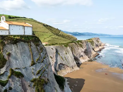 Ermita blanca sobre acantilados en la playa de Itzurun, con el mar Cantábrico y olas rompiendo.
