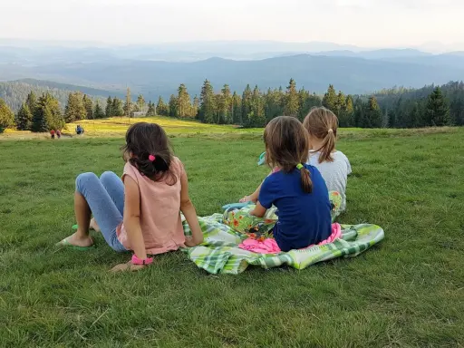 Trzy dziewczynki odpoczywają na kocu na trawie, podziwiając widok gór. W tle widać ludzi idących szlakiem.