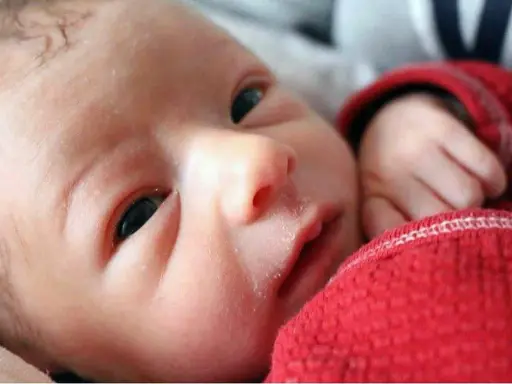 Primo piano di un neonato con lieve crosta lattea sulle sopracciglia, avvolto in una coperta rossa.