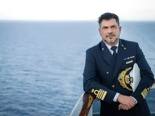 Il comandante MSC Splendida, con la sua uniforme blu scuro e il cappello dorato, posa con il mare calmo sullo sfondo.