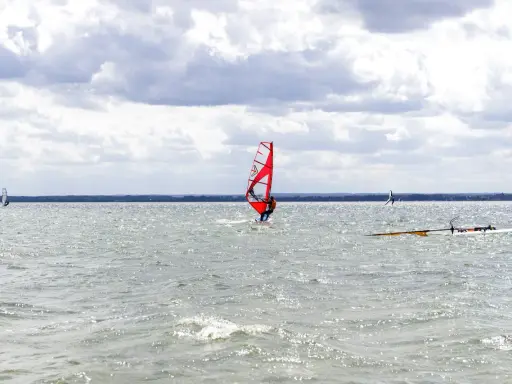 Czerwony żagiel do windsurfingu na wodzie, surfer w akcji. W tle inni windsurferzy i chmury.