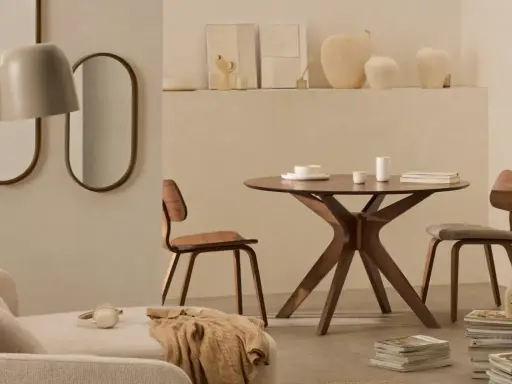 Dos espejos ovalados cuelgan sobre un sofá beige. A la derecha, una mesa redonda de madera con sillas y un jarrón.