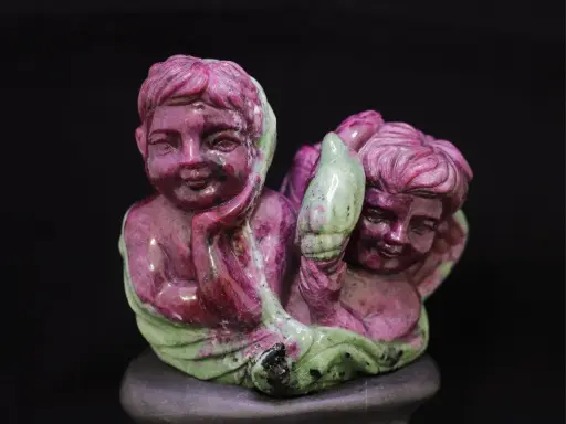 Due putti scolpiti in pietra, uno con la mano sul mento, sembrano abbracciare un elemento verde, forse un fiore. La pietra, un rubino verde, dona un tocco di magia.