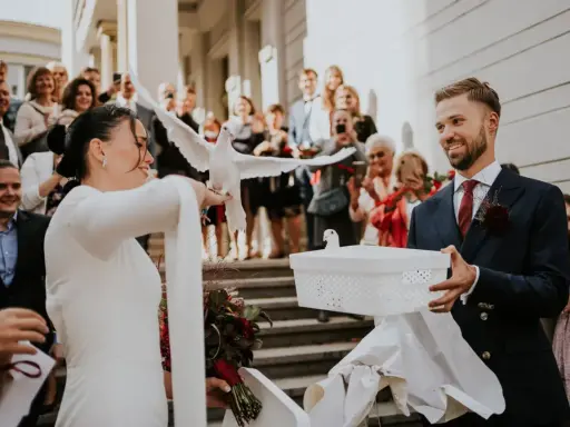 Para młoda wypuszcza gołębie na kameralnym weselu. W tle widać gości z telefonami.