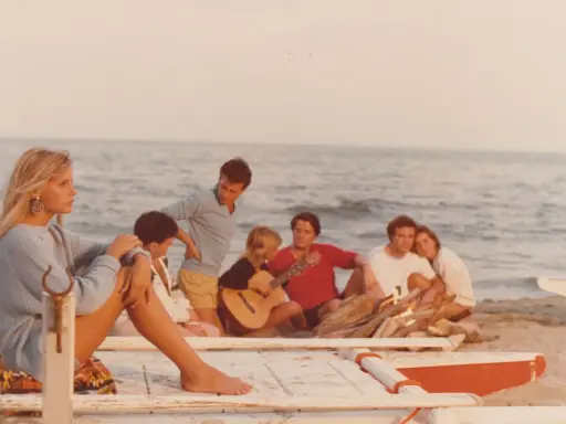 Giovani su una spiaggia, un gruppo di amici che ricorda i film estivi italiani, con chitarra e falò.