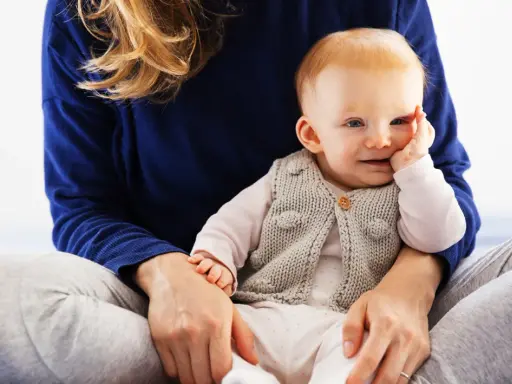 Uśmiechnięte dziecko w sweterku, które zaczyna siadać, siedzi na kolanach mamy.