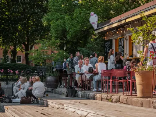 Folk njuter av solen på uteserveringar i Stockholm. En glasskylt syns ovanför en servering.
