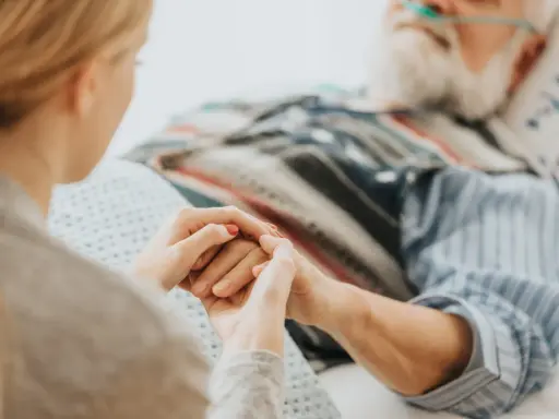Mani che si stringono tra una giovane donna e un anziano a letto, con cannule nasali. Il caregiver viene pagato per la sua dedizione.