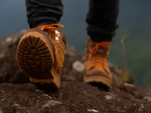 Stopy w brązowych butach trekkingowych z pomarańczowymi sznurówkami na kamienistym szlaku. Membrana w butach zapewnia komfort podczas wędrówki.