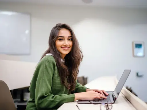 Uśmiechnięta studentka pracuje na laptopie w sali wykładowej. Jej status ucznia jest widoczny w otoczeniu i skupieniu na nauce.